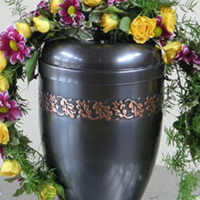 Schwarze Urne mit goldener Verzierung, umgeben von gelbem und lila Blumenschmuck.